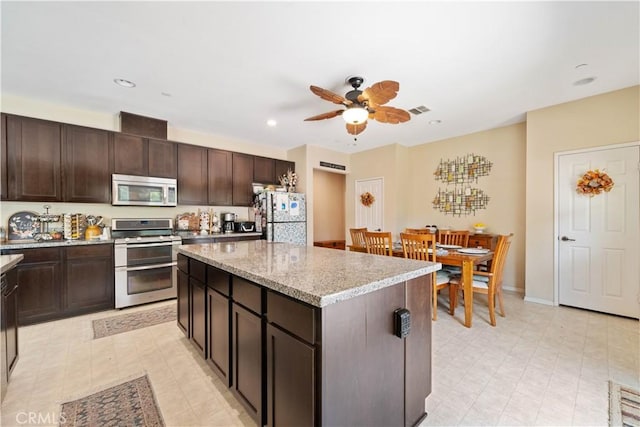 kitchen with stainless steel appliances, a center island, light stone counters, ceiling fan, and dark brown cabinets