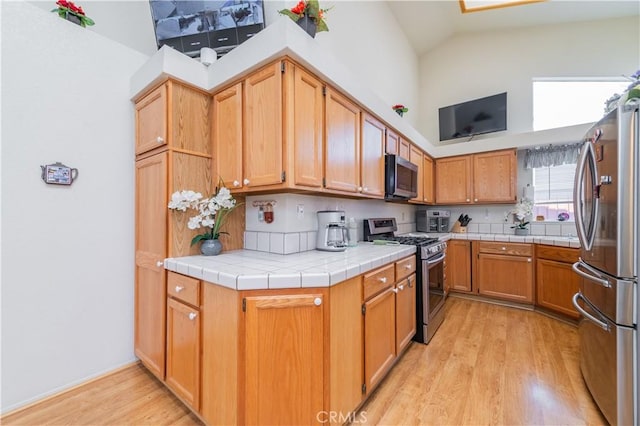 kitchen featuring lofted ceiling with skylight, light hardwood / wood-style flooring, appliances with stainless steel finishes, tile counters, and kitchen peninsula