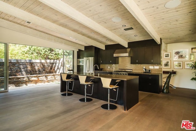 kitchen with hardwood / wood-style floors, a center island with sink, a kitchen breakfast bar, wall chimney range hood, and beamed ceiling