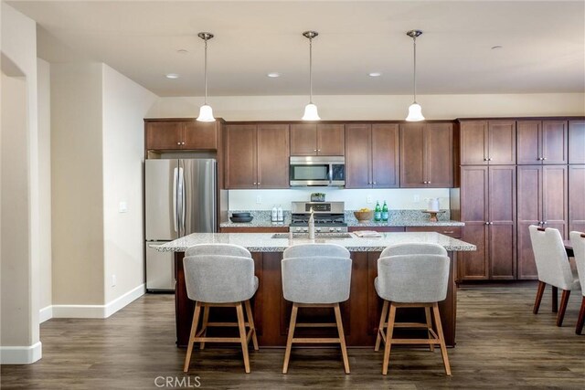 kitchen featuring an island with sink, dark wood finished floors, appliances with stainless steel finishes, baseboards, and hanging light fixtures