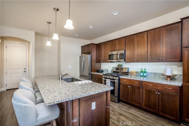 kitchen with sink, hanging light fixtures, stainless steel appliances, light stone counters, and an island with sink