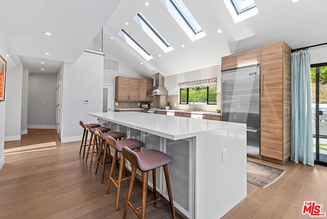 kitchen with a large island, stainless steel built in refrigerator, wall chimney range hood, high vaulted ceiling, and a skylight