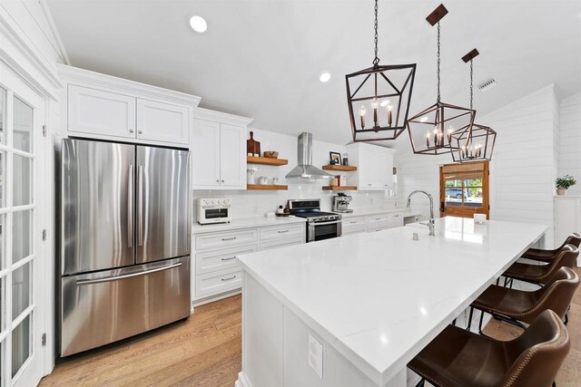 kitchen with a breakfast bar, hanging light fixtures, wall chimney exhaust hood, white cabinetry, and stainless steel appliances