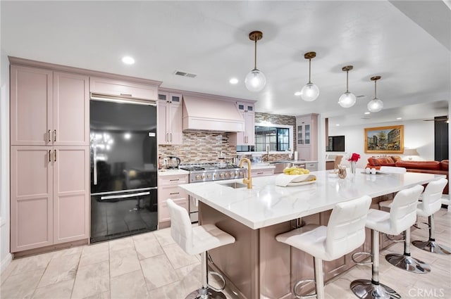 kitchen with hanging light fixtures, a center island with sink, light stone counters, black refrigerator, and premium range hood