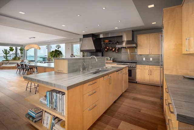 kitchen with sink, a kitchen island with sink, high end stove, wall chimney exhaust hood, and light brown cabinets