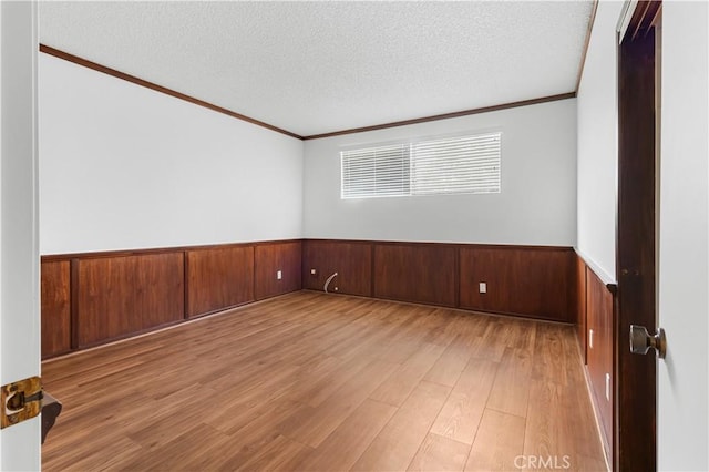 unfurnished room with a textured ceiling, wooden walls, light wood-style floors, wainscoting, and crown molding