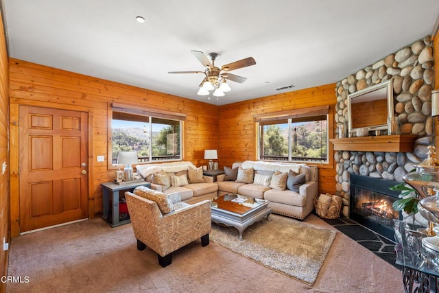 carpeted living room featuring wood walls, ceiling fan, plenty of natural light, and a stone fireplace