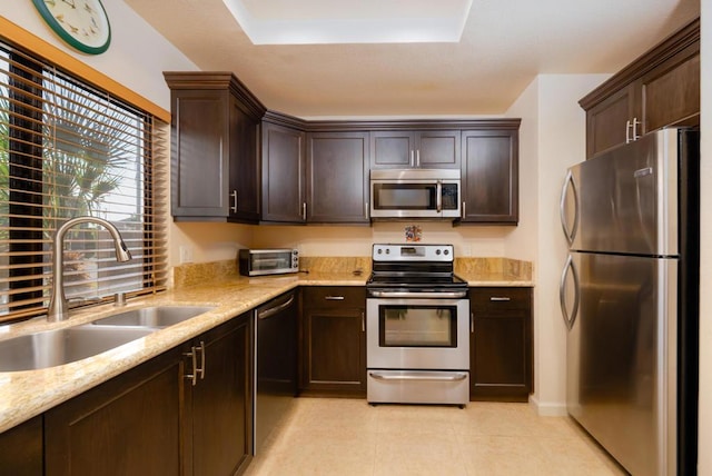 kitchen with sink, light stone countertops, a tray ceiling, dark brown cabinets, and stainless steel appliances