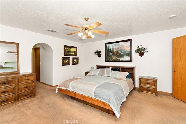 bedroom featuring light carpet, a textured ceiling, a ceiling fan, visible vents, and arched walkways