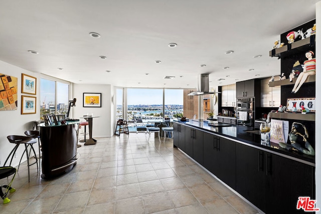 kitchen with light tile patterned floors, a breakfast bar, multiple ovens, island range hood, and expansive windows