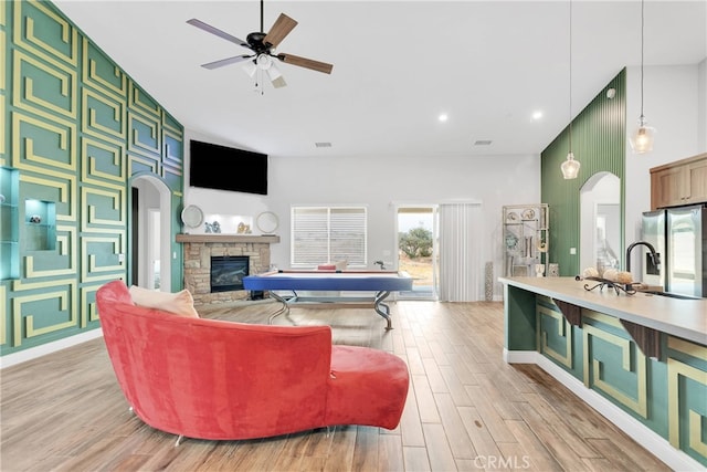 living room featuring light wood-type flooring, sink, a stone fireplace, a high ceiling, and ceiling fan