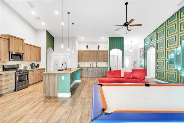 kitchen with a towering ceiling, stainless steel appliances, light hardwood / wood-style flooring, ceiling fan, and a kitchen island with sink