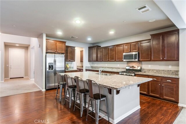 kitchen featuring a center island with sink, stainless steel appliances, a kitchen breakfast bar, light stone countertops, and dark hardwood / wood-style flooring