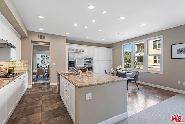 kitchen with appliances with stainless steel finishes, tasteful backsplash, light stone counters, a center island with sink, and white cabinetry