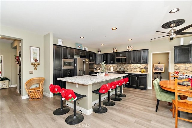 kitchen with a center island with sink, light wood-type flooring, light stone countertops, appliances with stainless steel finishes, and tasteful backsplash