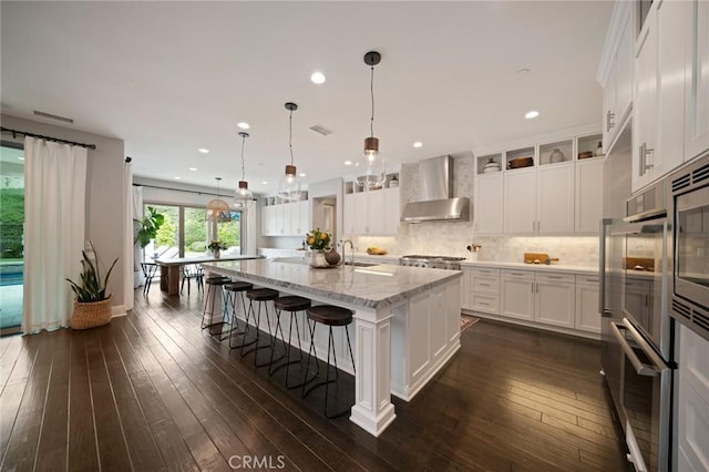 kitchen with a kitchen island with sink, dark wood-type flooring, white cabinets, wall chimney exhaust hood, and appliances with stainless steel finishes