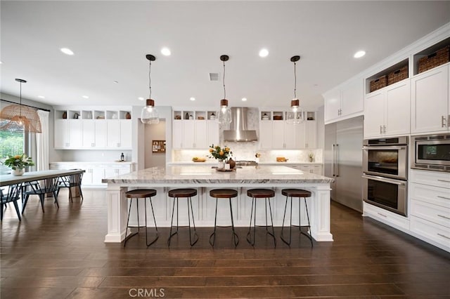 kitchen featuring white cabinets, a spacious island, built in appliances, wall chimney exhaust hood, and decorative light fixtures