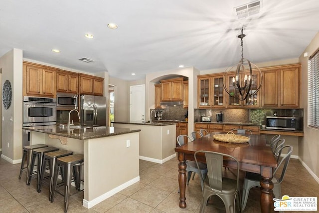 kitchen featuring appliances with stainless steel finishes, dark stone countertops, pendant lighting, a center island with sink, and a notable chandelier