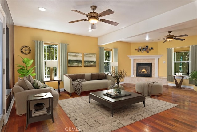 living room featuring beam ceiling, wood-type flooring, a tiled fireplace, and ceiling fan