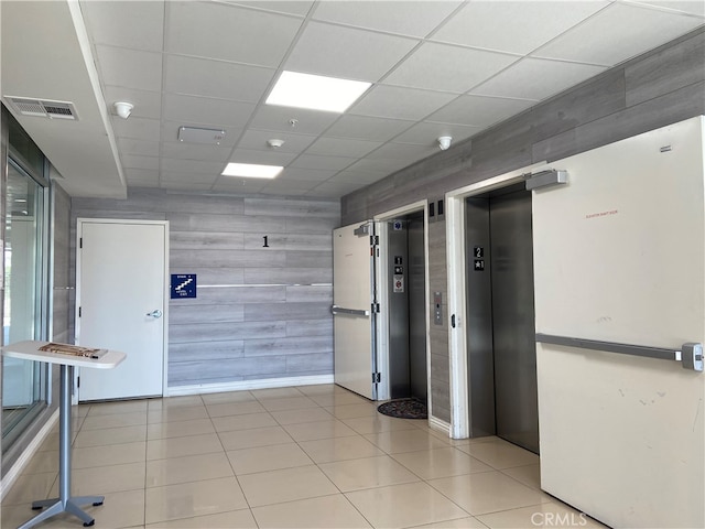 empty room featuring light tile patterned floors, a drop ceiling, wooden walls, and elevator