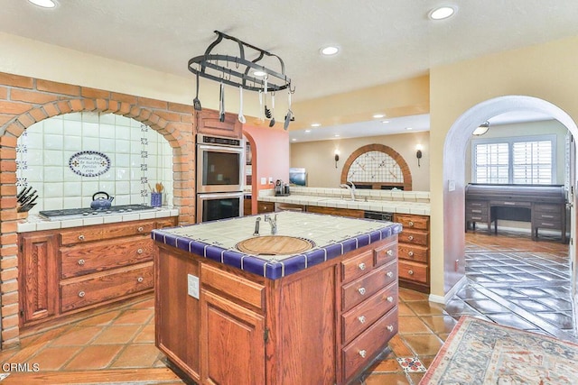 kitchen with tile countertops, backsplash, hanging light fixtures, an island with sink, and appliances with stainless steel finishes