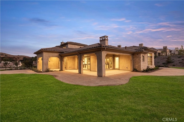 back house at dusk featuring a lawn and a patio area