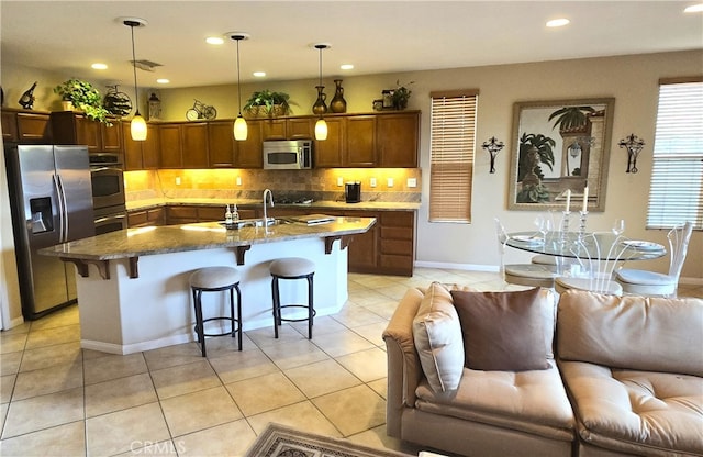 kitchen with light tile patterned floors, appliances with stainless steel finishes, an island with sink, decorative light fixtures, and a breakfast bar