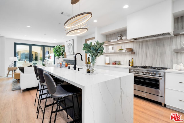 kitchen featuring light stone counters, light hardwood / wood-style floors, white cabinetry, a center island with sink, and high end stainless steel range