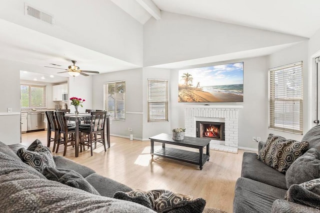 living room with ceiling fan, beamed ceiling, a brick fireplace, high vaulted ceiling, and light wood-type flooring