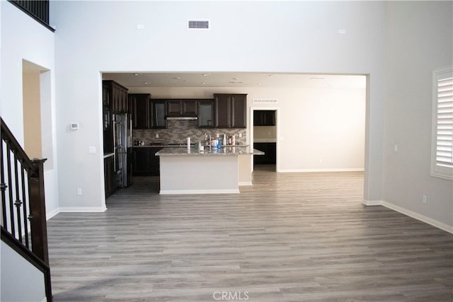 kitchen with dark brown cabinetry, an island with sink, backsplash, light wood-type flooring, and a towering ceiling