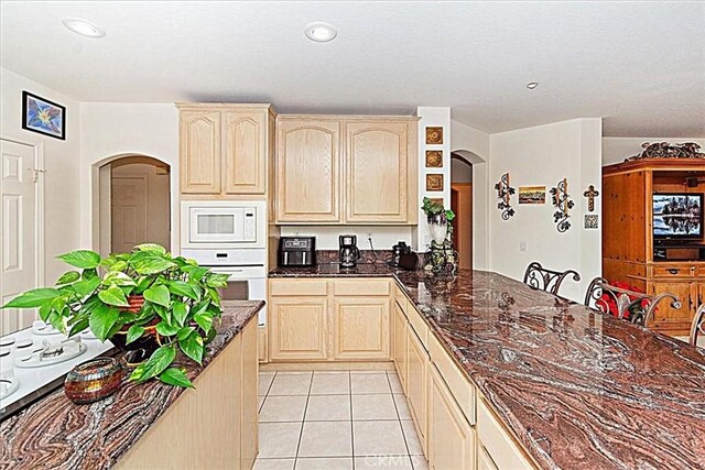 kitchen featuring light brown cabinets, light tile patterned floors, dark stone counters, and white appliances