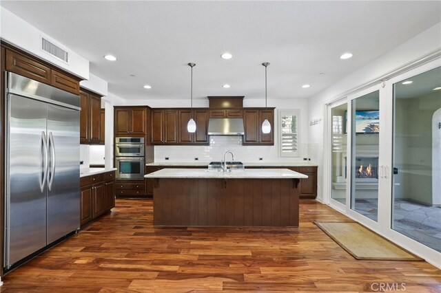 kitchen with decorative backsplash, stainless steel appliances, dark wood-type flooring, pendant lighting, and a center island with sink