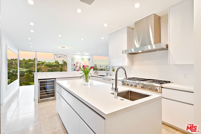 kitchen with sink, beverage cooler, white cabinets, wall chimney range hood, and a center island with sink