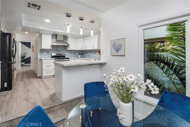 kitchen with kitchen peninsula, wall chimney exhaust hood, stainless steel appliances, pendant lighting, and white cabinetry