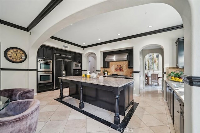 kitchen featuring a kitchen island, built in appliances, ventilation hood, light stone counters, and crown molding