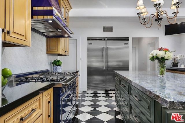 kitchen with dark stone countertops, black gas range oven, a chandelier, wall chimney range hood, and stainless steel built in fridge