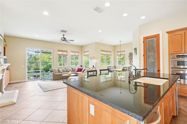 kitchen with a kitchen island with sink, sink, ceiling fan, dark stone countertops, and stainless steel double oven