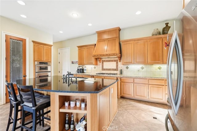 kitchen with backsplash, a kitchen island with sink, light tile patterned floors, appliances with stainless steel finishes, and custom range hood