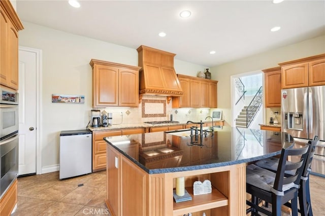 kitchen with tasteful backsplash, an island with sink, dark stone counters, custom range hood, and appliances with stainless steel finishes