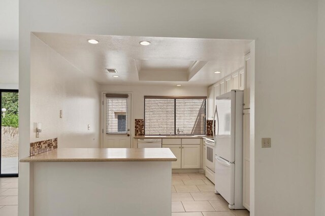 kitchen with kitchen peninsula, white appliances, a tray ceiling, light tile patterned floors, and white cabinetry