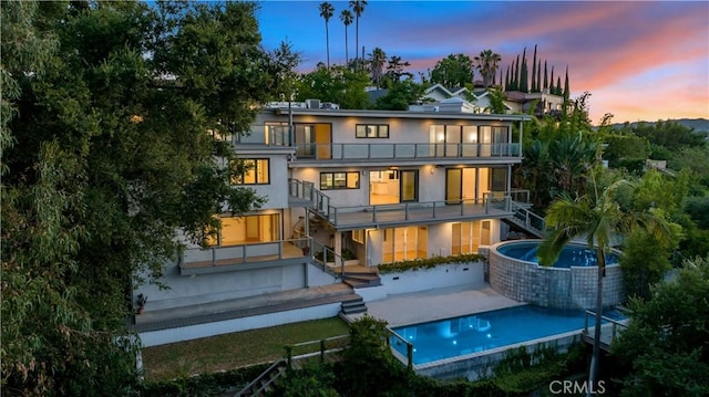 back of property at dusk featuring stucco siding, stairway, a balcony, an in ground hot tub, and a patio area