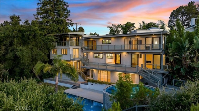 back of house at dusk featuring stucco siding, a patio, a balcony, and stairs