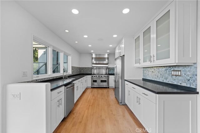 kitchen featuring a sink, stainless steel appliances, white cabinetry, dark countertops, and wall chimney range hood