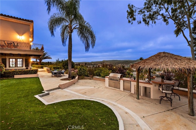 patio terrace at dusk with a grill, a balcony, a yard, and exterior kitchen