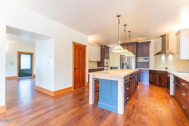 kitchen featuring a kitchen island with sink, stainless steel appliances, light countertops, wall chimney exhaust hood, and decorative light fixtures