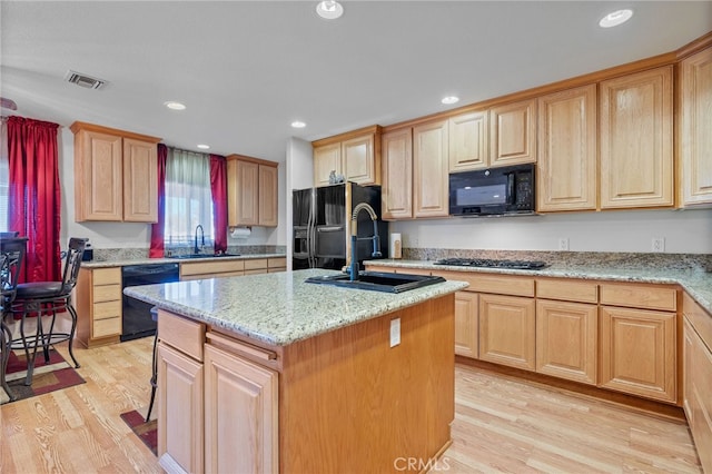 kitchen featuring light wood-type flooring, sink, an island with sink, black appliances, and light stone countertops