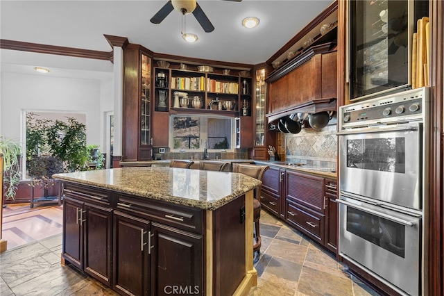 kitchen featuring light stone counters, ornamental molding, a kitchen island, backsplash, and double oven