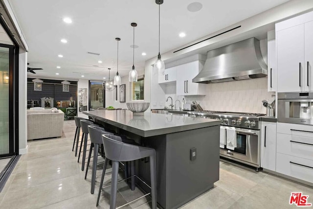 kitchen featuring pendant lighting, wall chimney range hood, white cabinetry, appliances with stainless steel finishes, and a kitchen bar