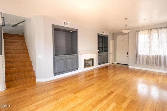 unfurnished living room featuring visible vents, baseboards, light wood-style floors, stairway, and a glass covered fireplace