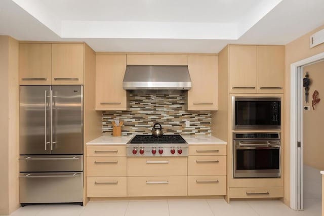 kitchen with wall chimney range hood, backsplash, built in appliances, a tray ceiling, and light brown cabinetry